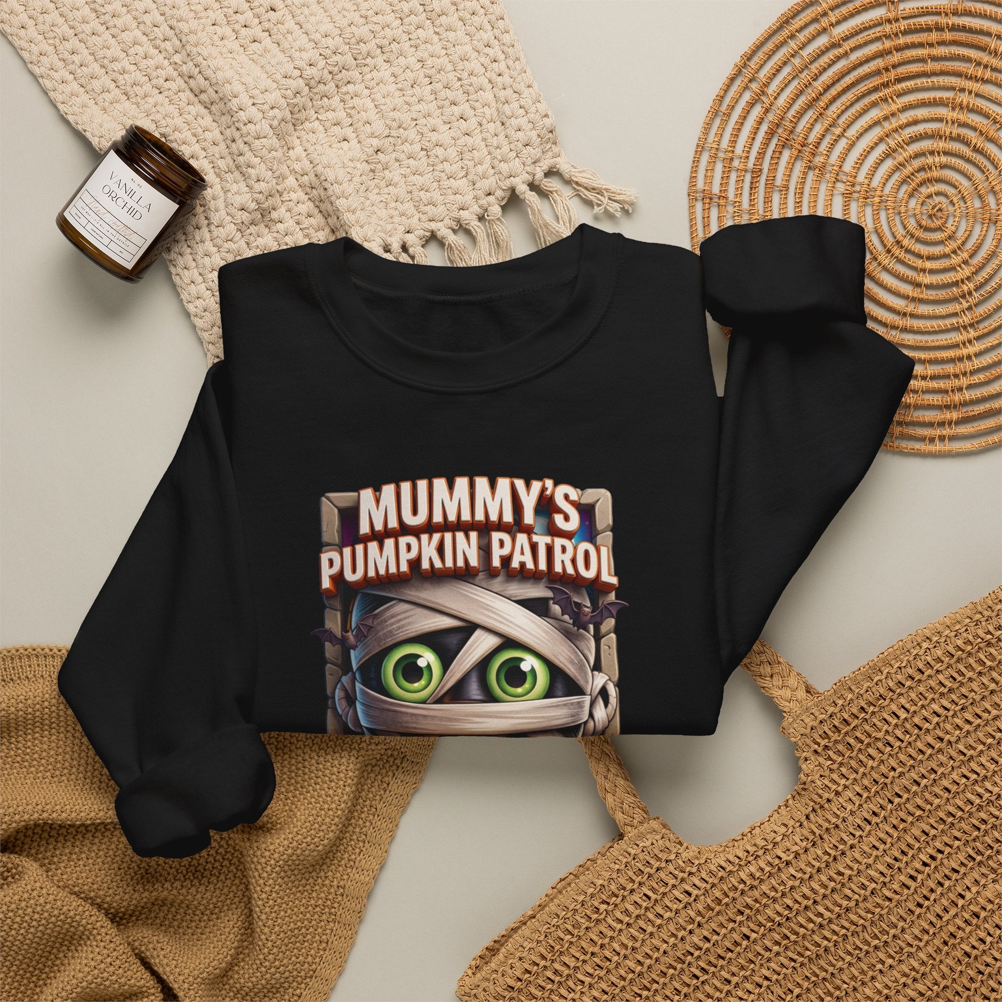 Black crewneck sweatshirt styled flat with a candle — cartoon mummy hugging a carved pumpkin