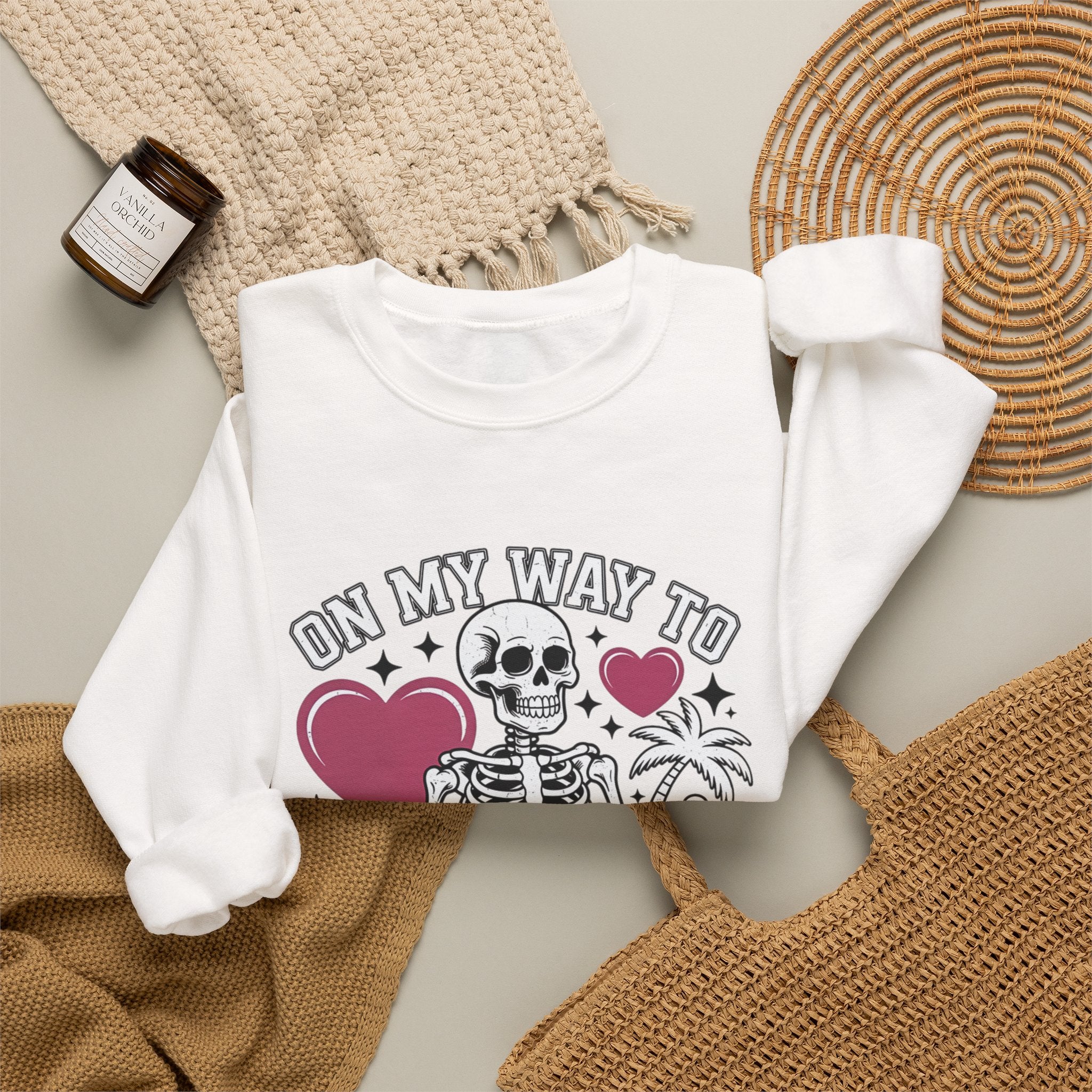 White crewneck sweatshirt styled flat with a candle — line-drawn skeletons under pink hearts